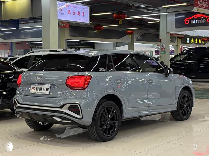 Задний ракурс серой Audi Q2L 35 TFSI в автосалоне: видны задняя оптика, бампер, тёмные диски и дилерские стенды на фоне.