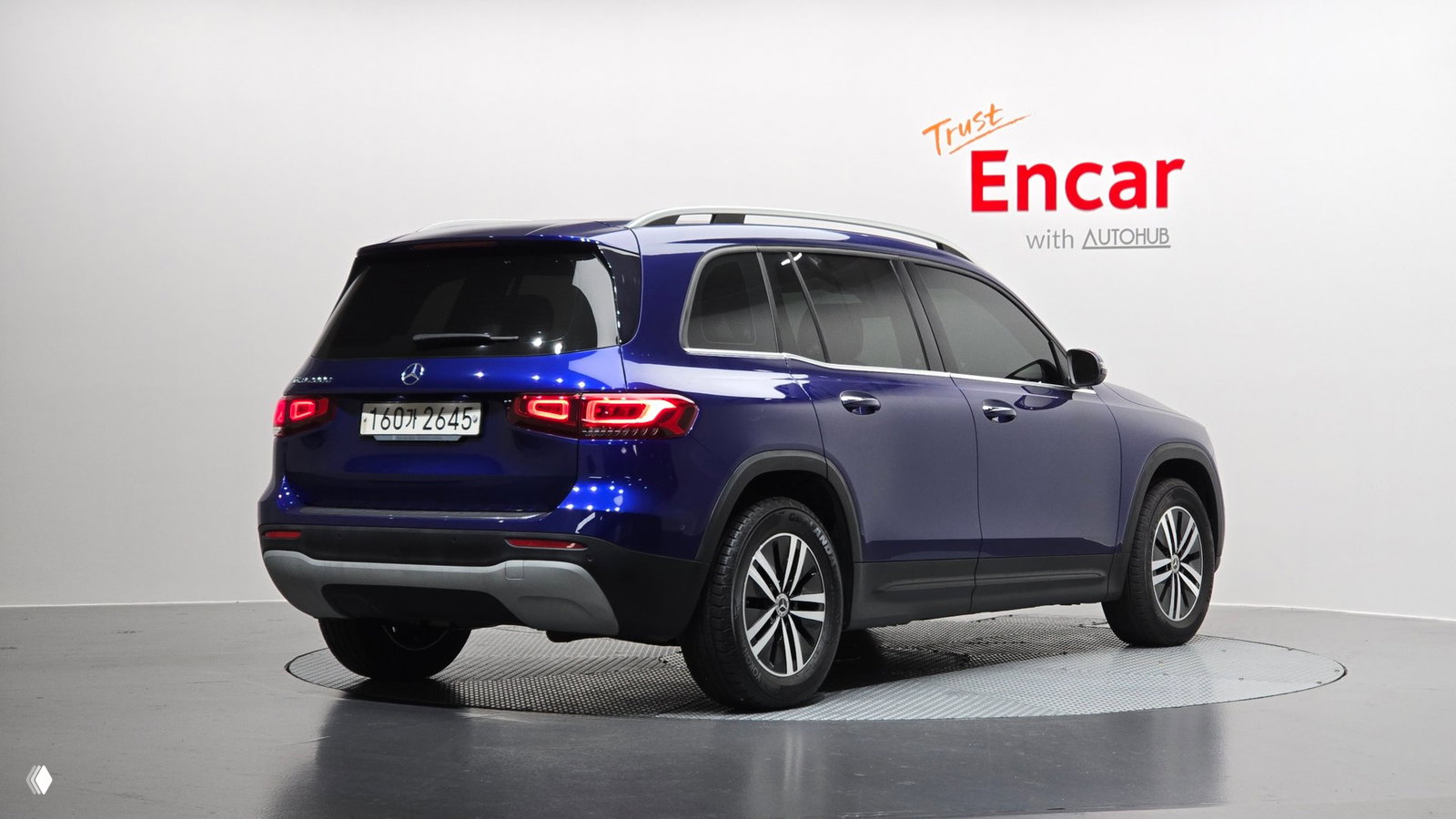 Задний и боковой ракурс синего Mercedes GLB на студийной платформе, видны задние фонари, форма багажника и установленный номерной знак.