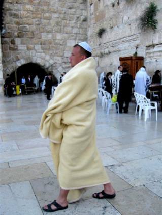 Паломник, завернутый в белое покрывало у Стены Плача, идёт в сандалиях мимо пластиковых стульев и других молящихся.