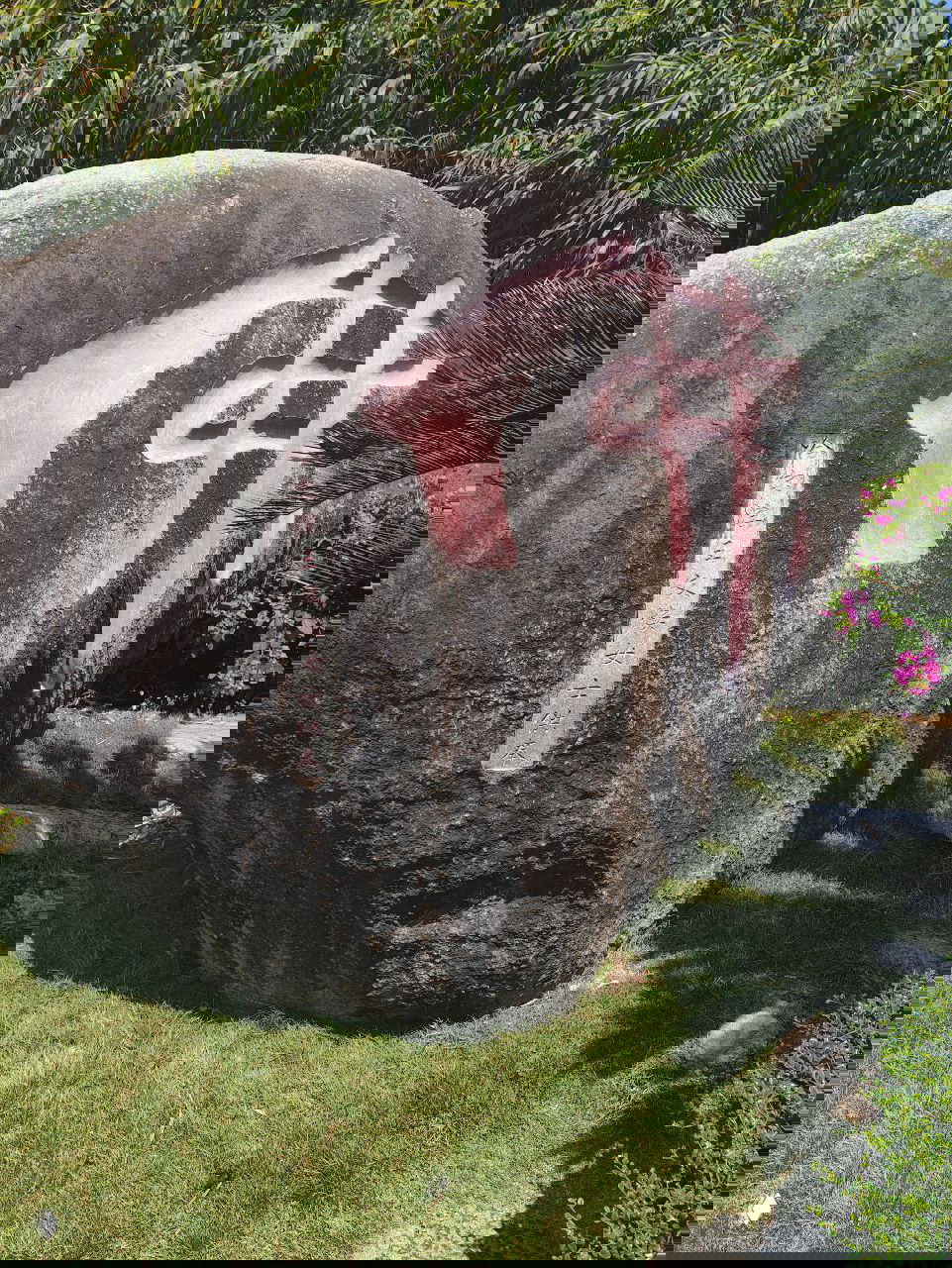Большой природный валун с нанесённым крупным китайским иероглифом на солнечном участке парка, вокруг видна зелень и цветы.