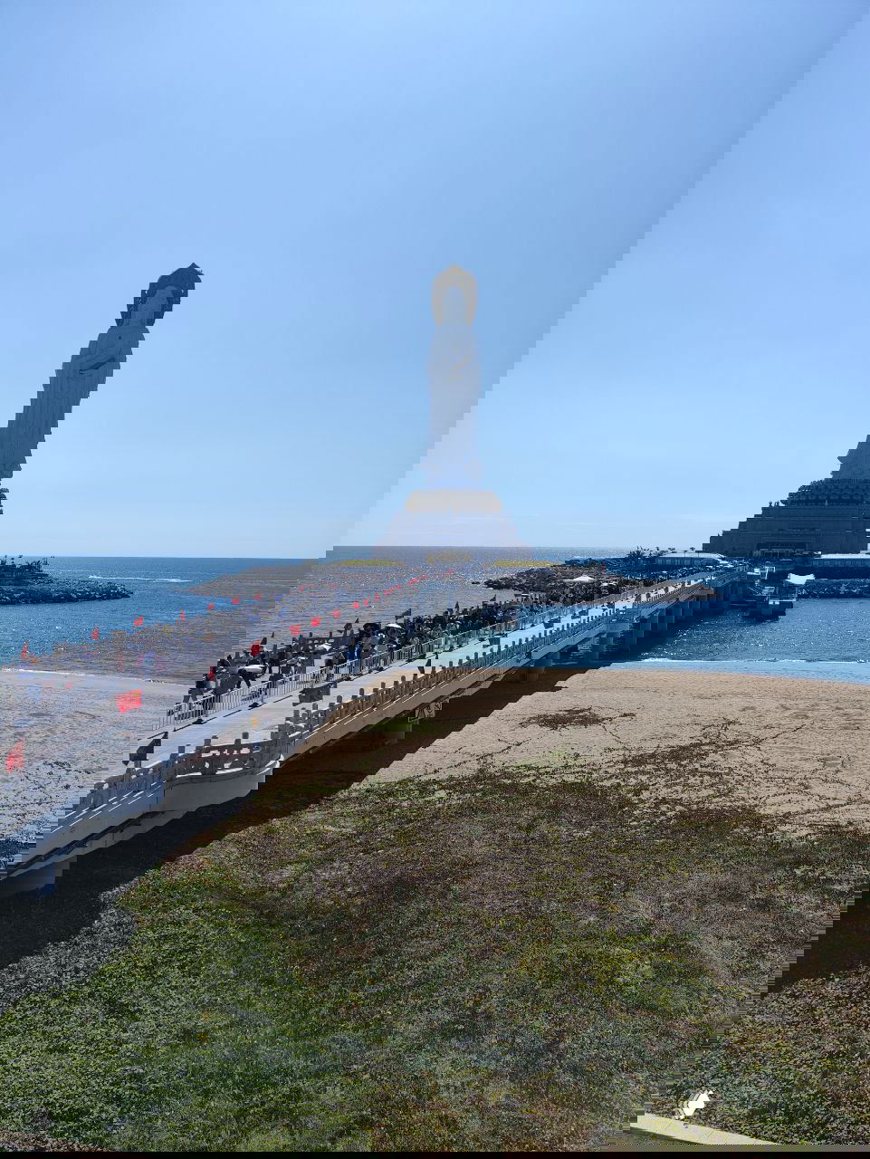 Высокая прибрежная статуя на искусственном мысе; к памятнику ведёт пешеходный мост, вокруг море и множество туристов на набережной.