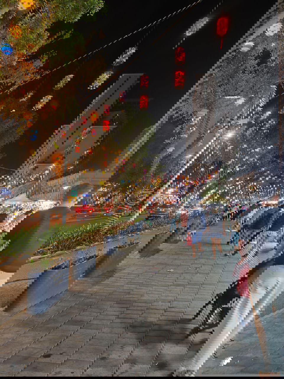 Ночная городская улица украшена бумажными фонариками; прохожие идут мимо уличных лотков и витрин на пешеходной зоне рынка.