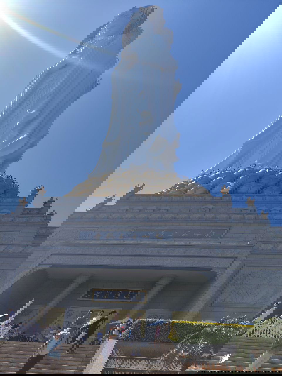 Боковой вид гигантской статуи Гуань Инь с золотым лотосовым пьедесталом, лестницей и туристами на переднем плане под ясным небом.