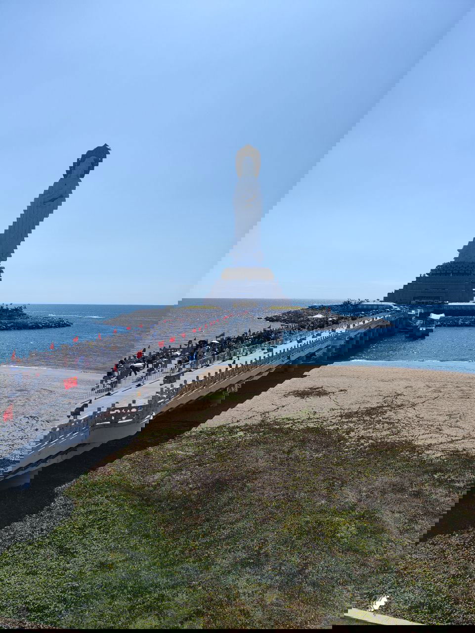 Длинная набережная и пешеходный мост к островной статуе Гуань Инь; туристы идут по пирсу, по сторонам видна морская гладь и флаги.