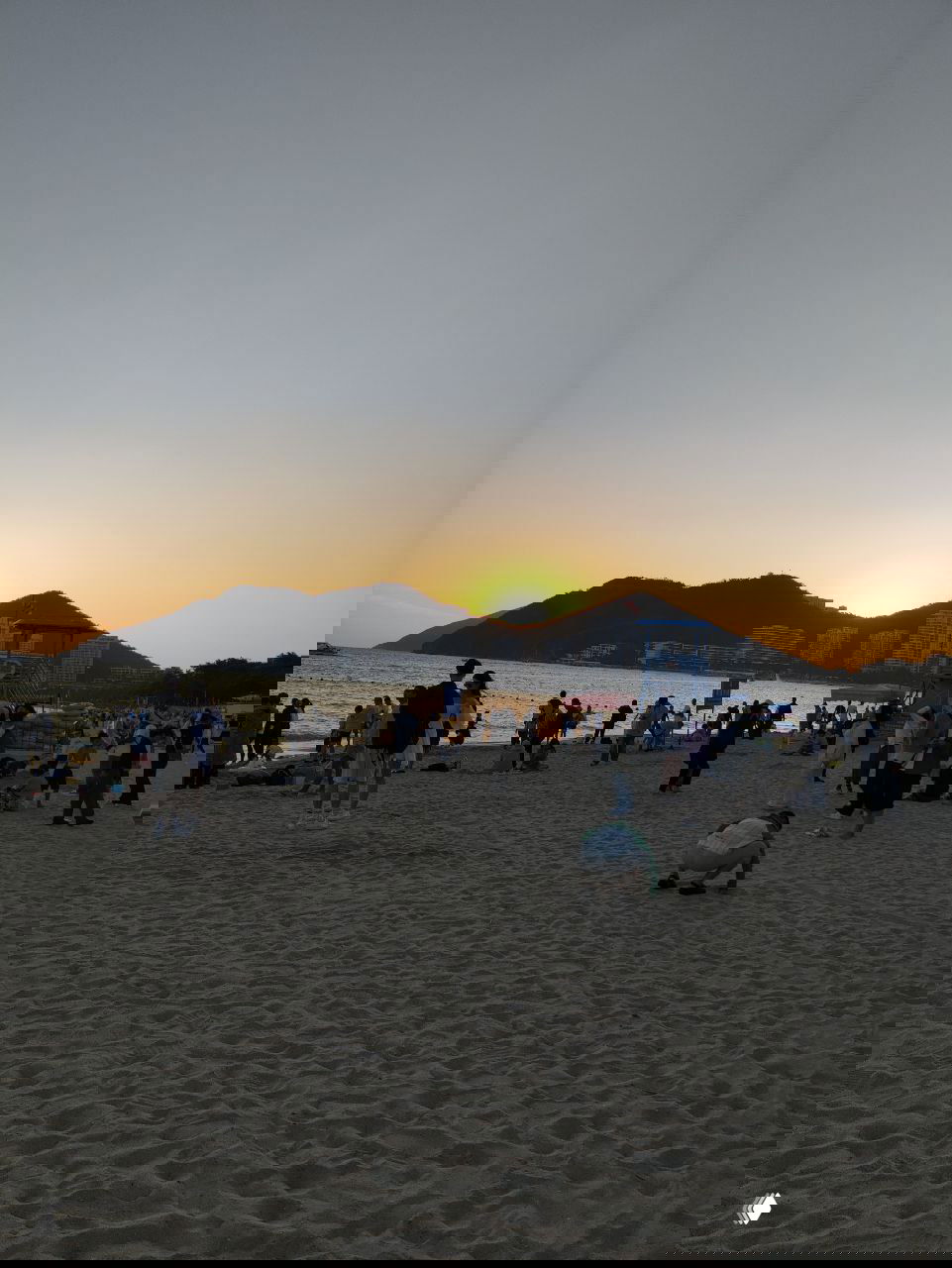 Пляжный закат: много отдыхающих на берегу, силуэты людей на фоне заходящего солнца и тёмные контуры холмов на горизонте.