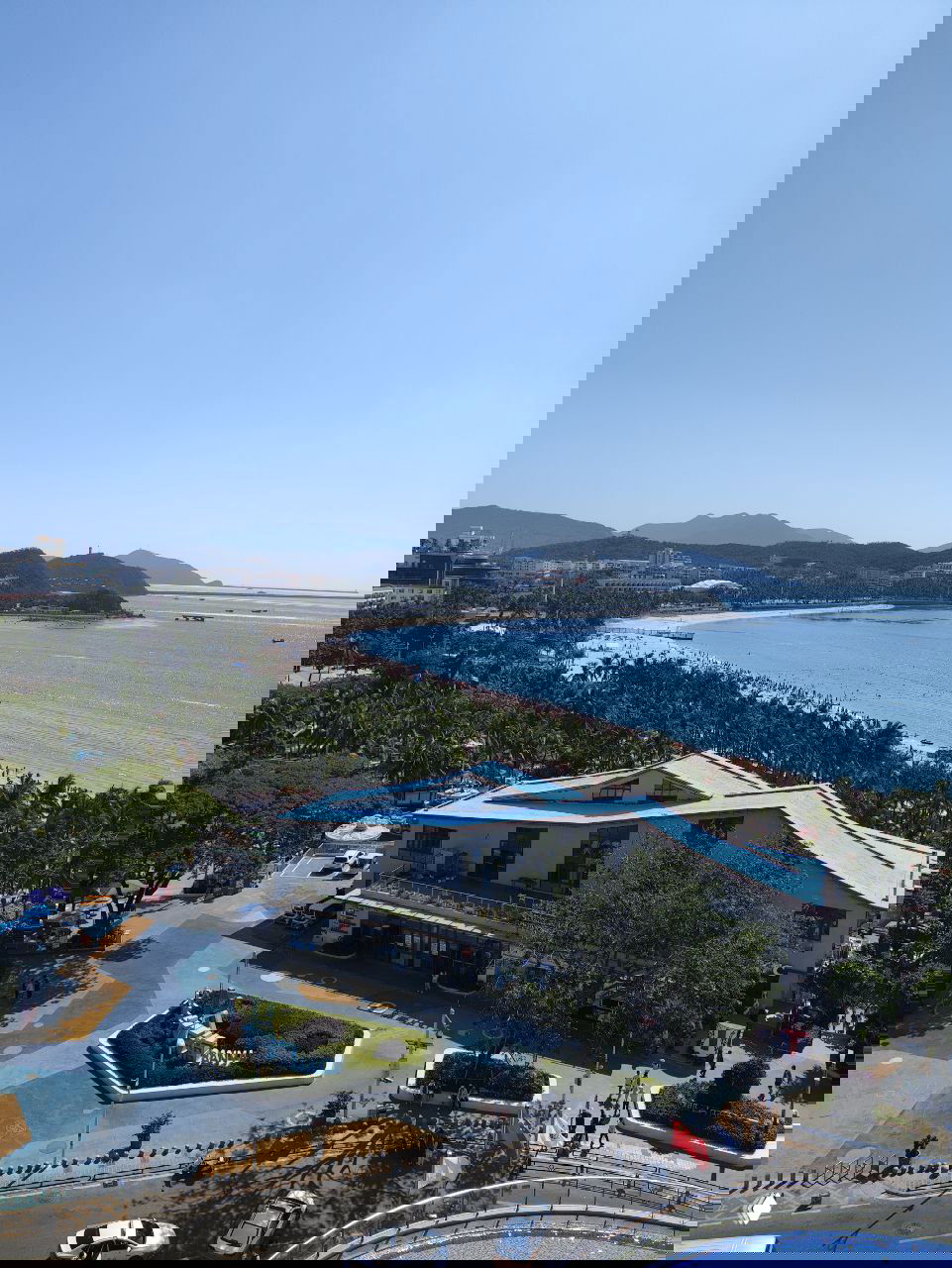 Прибрежный вид с пляжем и зданием с голубой крышей; пальмы, населённая береговая линия и инфраструктура туристической зоны на переднем плане.