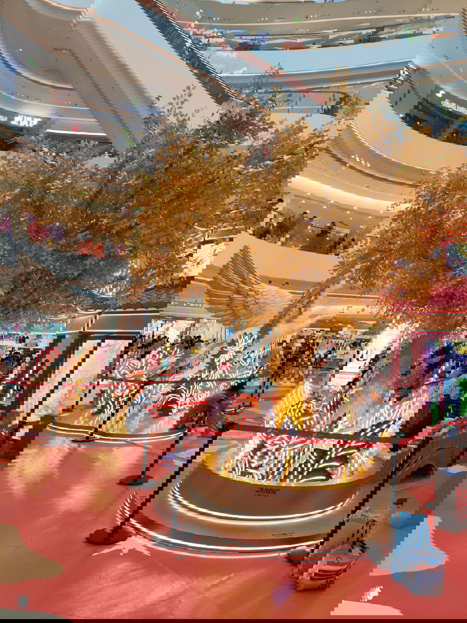 Интерьер торгового центра с большой золотой декоративной «деревянной» инсталляцией и светящимися цветами, люди гуляют вокруг украшения.