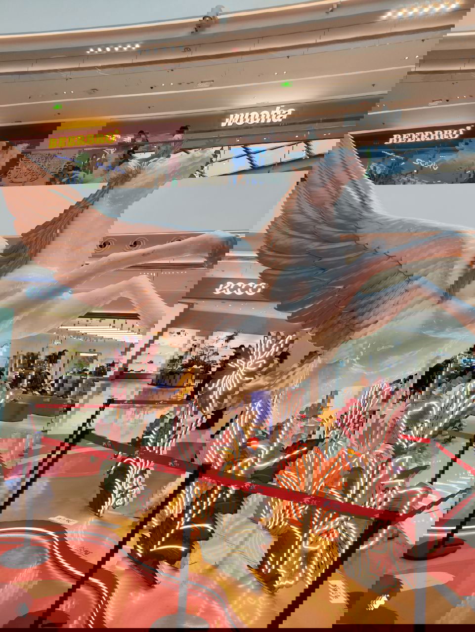 Белая статуя пегаса с расправленными крыльями в торговом центре, окружённая неоновыми цветочными декорациями и ограждениями для посетителей.