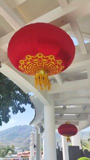 Крупный красный декоративный китайский фонарь с золотым орнаментом, подвешенный под сводом в общественном пространстве на фоне гор и неба.