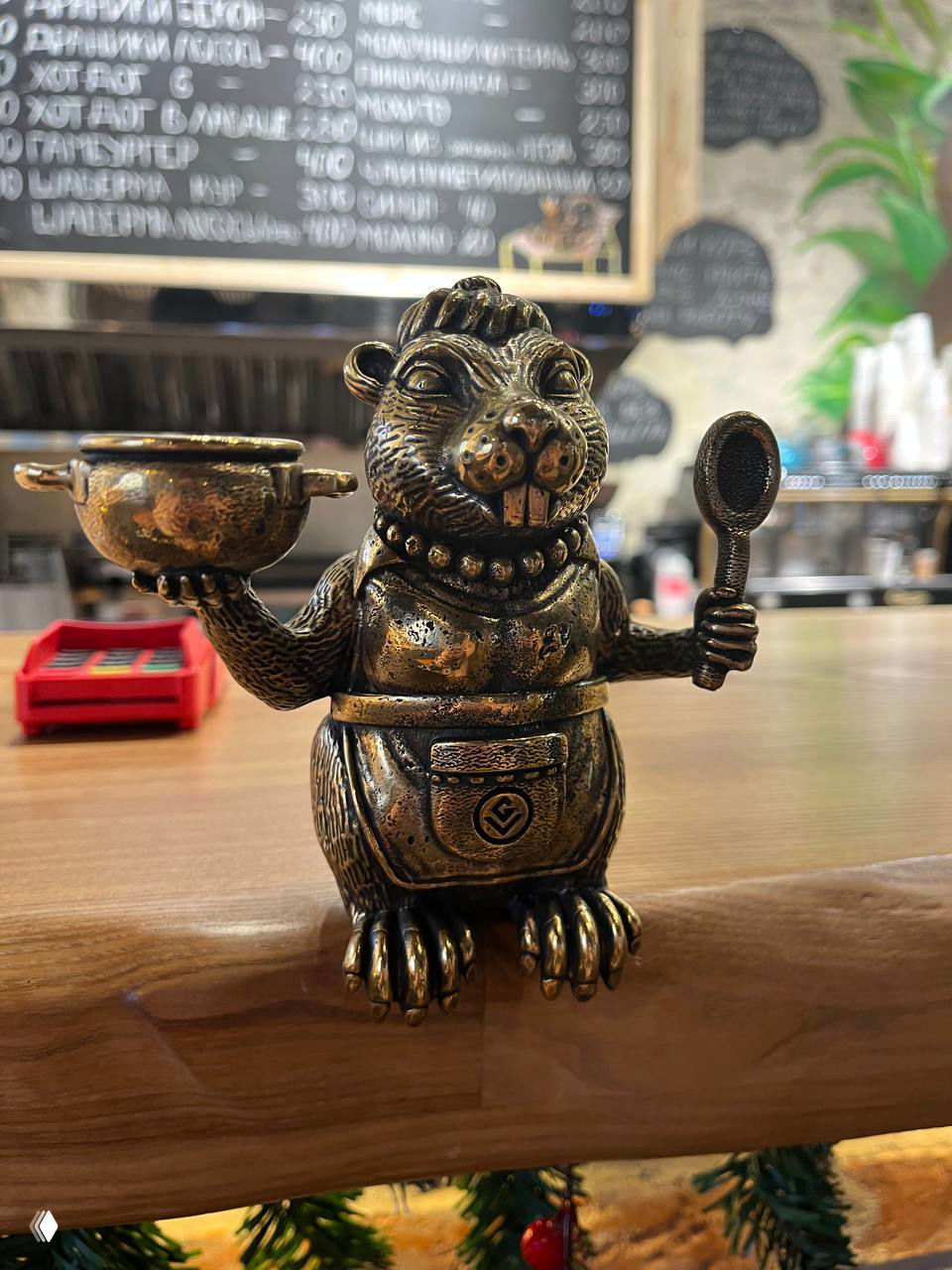 Бронзовая фигурка бобра в фартуке держит ложку и кастрюлю на прилавке кафе, на заднем плане меню и кухня.