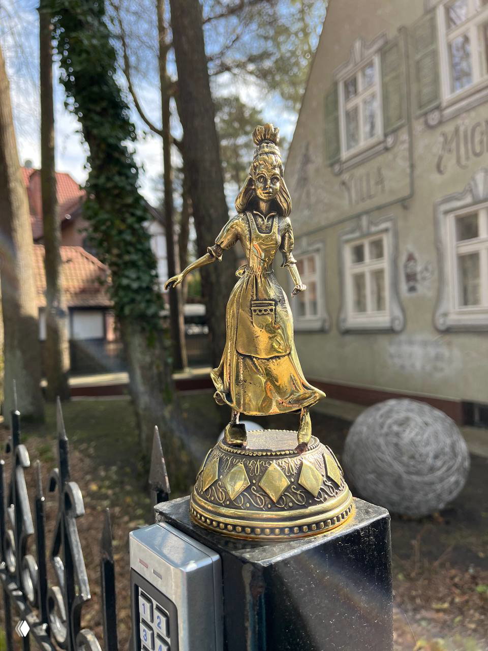 Золотистая фигурка девочки на металлической колонне у кованого забора на фоне соснового сквера и старого дома в Светлогорске