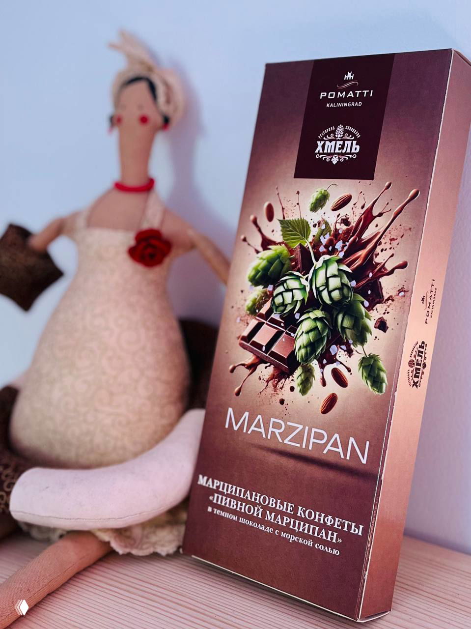 Коробка пивного марципана Pomatti с изображением шишек хмеля и плиток тёмного шоколада на фоне текстильной куклы