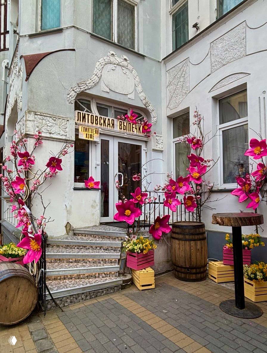 Украшенный вход в пекарню «Литовская выпечка» в Зеленоградске: декоративные большие бумажные цветы, деревянные бочки и весенние горшки у ступенек фасада.