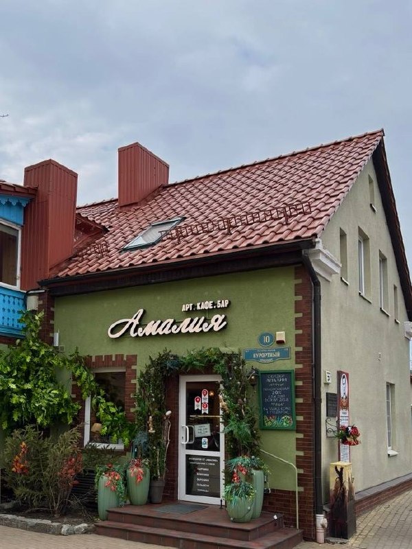 Фасад арт-кафе «Амалия» в Зеленоградске: зелёная вывеска, декоративные растения в горшках и уютный вход под красной черепичной крышей весной.