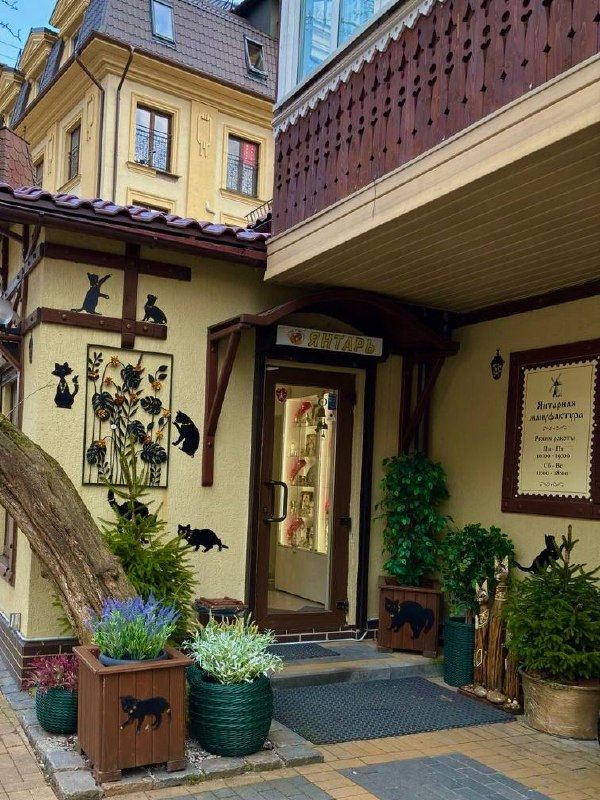 Вход в янтарную мануфактуру в Зеленоградске с силуэтами котов на фасаде, декоративными кашпо и зелеными растениями — уютный дворик в старой части города.
