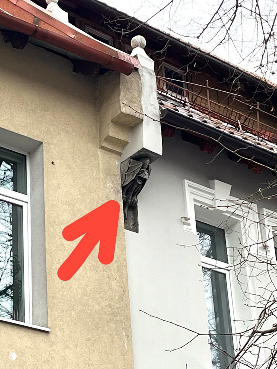 Крупный план горгульи под скатом крыши, отмеченной стрелкой; видна скульптурная деталь водостока на фоне стены здания.