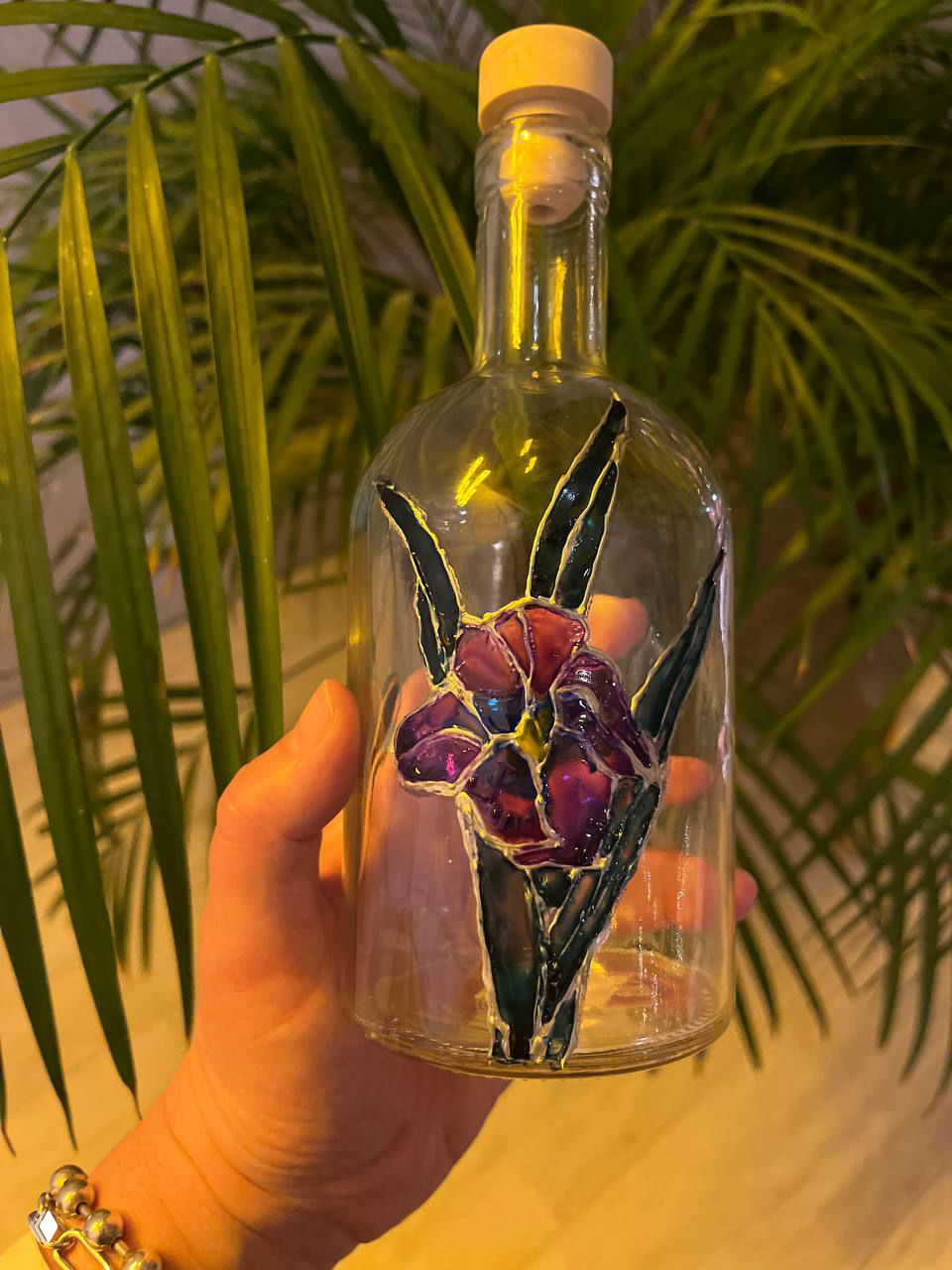 Готовый витраж на бутылке с цветком пурпурного и зелёного цветов, рука держит бутылку на фоне зелёного растения — финальный результат работы.