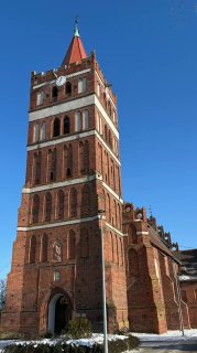 Красная кирпичная колокольня Свято-Георгиевского храма в Правдинске на фоне яркого синего неба; вид фасада с аркой входа, снежные остатки у основания, городской пейзаж.