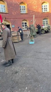 Сцена съёмок во дворе: актёры в исторических костюмах, реквизит — бочки и машина на фоне кирпичных фасадов.