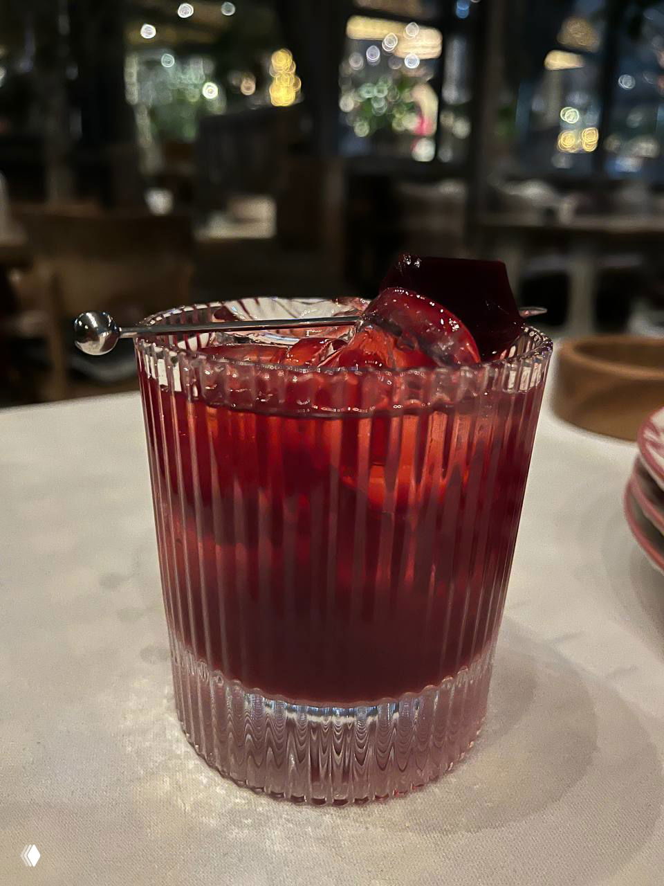 Коктейль — вишневый негрони в низком рифлёном бокале со льдом и вишнёвой гарнировкой, насыщенный рубиновый цвет на фоне ресторана.