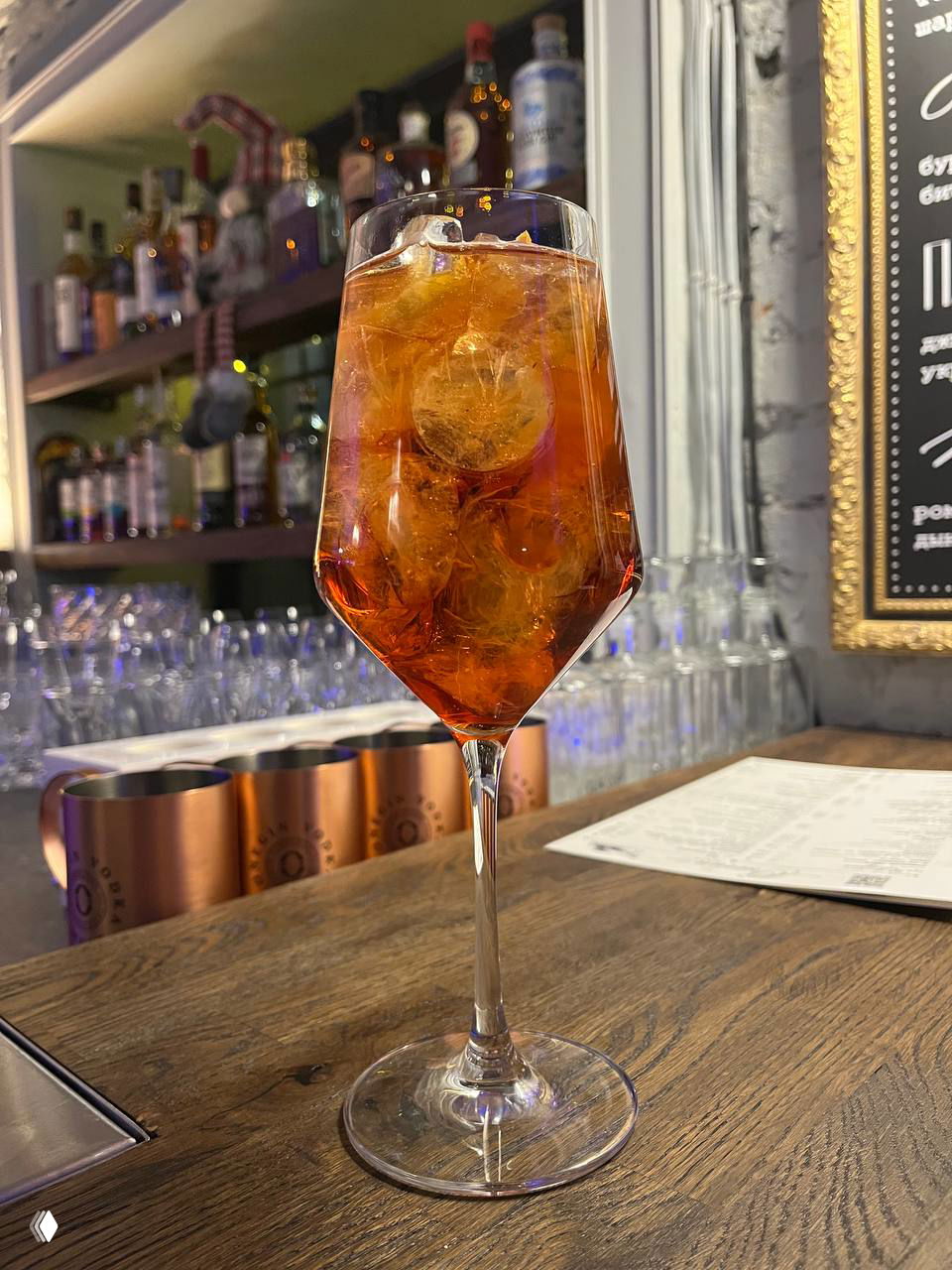 Бокал с ярким апельсиново-красным коктейлем на деревянной барной стойке, лёд и отражения подсветки, фон с полками и посудой бара Onegin Bar.