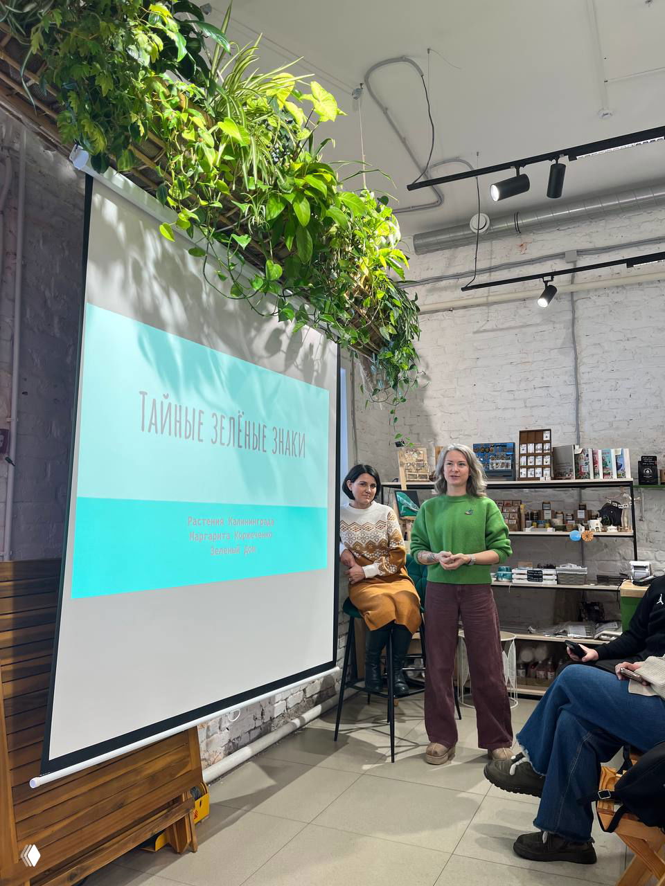 Проектор и сцена с презентацией «Тайные зелёные знаки» и два выступающих спикера в магазине, где проходит лекция про растения Калининграда