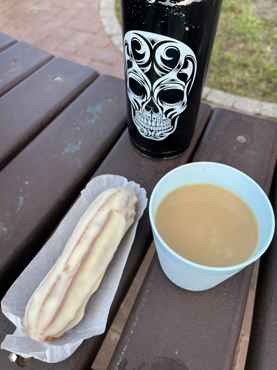 Эклер в белой глазури на бумажной подложке, бумажный стакан с кофе и термос с рисунком черепа на скамье в Железнодорожном районе.