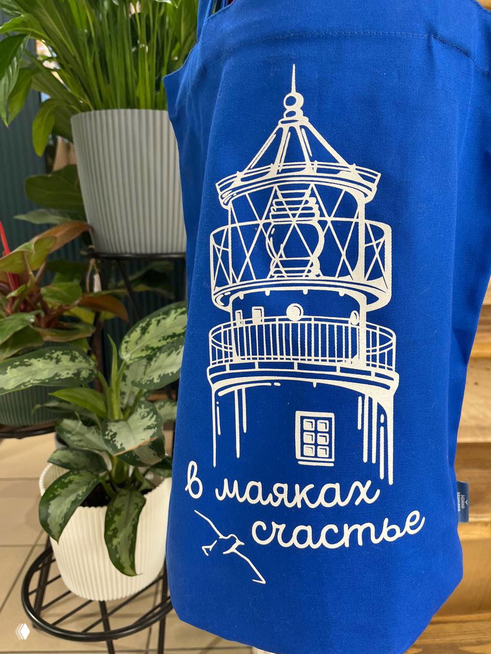 Фартук с принтом маяка и надписью «в маяках счастье», на фоне растений — локальный дизайнерский сувенир из магазина Калининграда