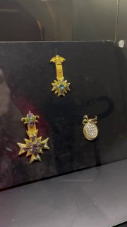 Крупный план нескольких декоративных брошей и кулонов на тёмном фоне экспозиции; видны эмаль, кристаллы и филигранные детали под светом.