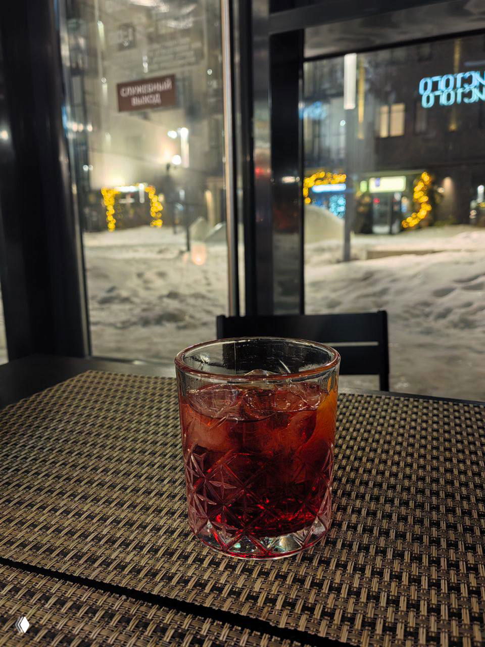 Стеклянный стакан с красным коктейлем и льдом на столе рюмочной, на заднем плане зимний городской пейзаж за витриной и приглушённое освещение.