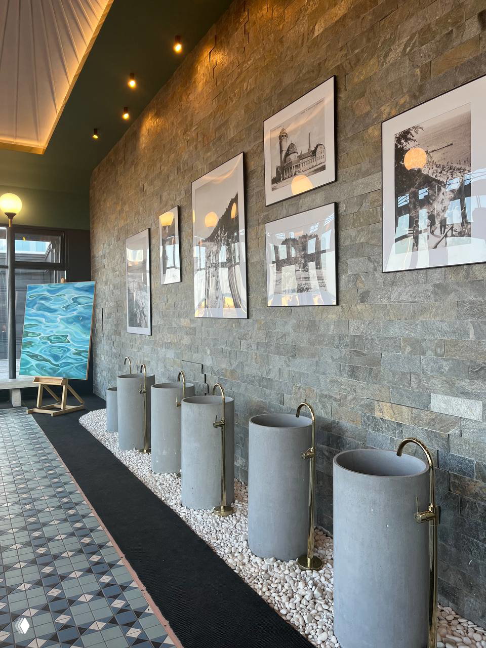 Интерьер бювета: ряд цилиндрических чаш с золотыми кранами на фоне каменной облицовки, настенные фотографии и узорная плитка на полу.