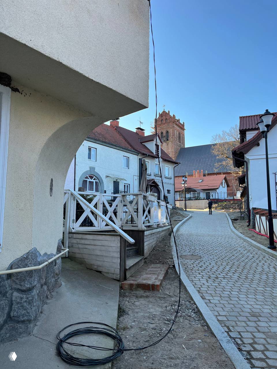 Улица в Калининграде: сбоку — закруглённый скос фасада дома, деревянные ограждения, мощёная дорога и кирха на фоне голубого неба.