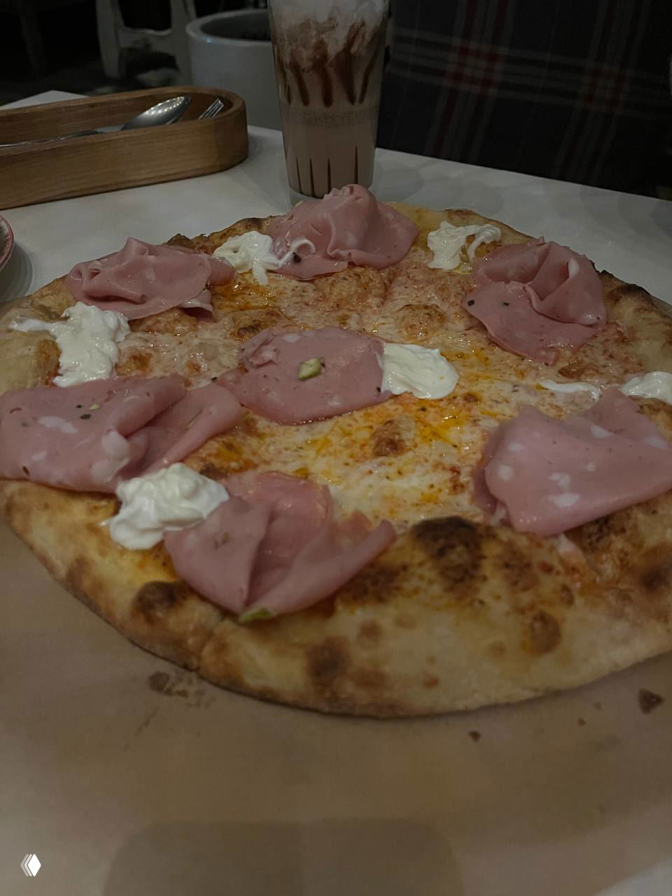 Пицца с ломтиками ветчины и кремовыми вставками на тонкой корочке, тёплая подача и дымок от дровяной печи на заднем плане.