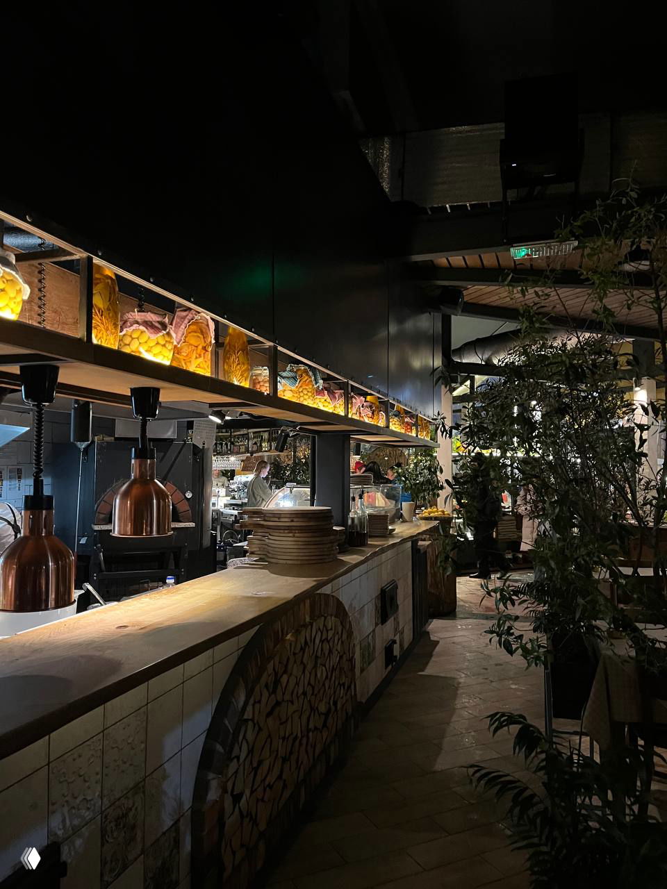 Открытая линия кухни с дровяной печью для пиццы и рабочей зоной шефа, вид на полки с ингредиентами и тёплое освещение внутри ресторана.