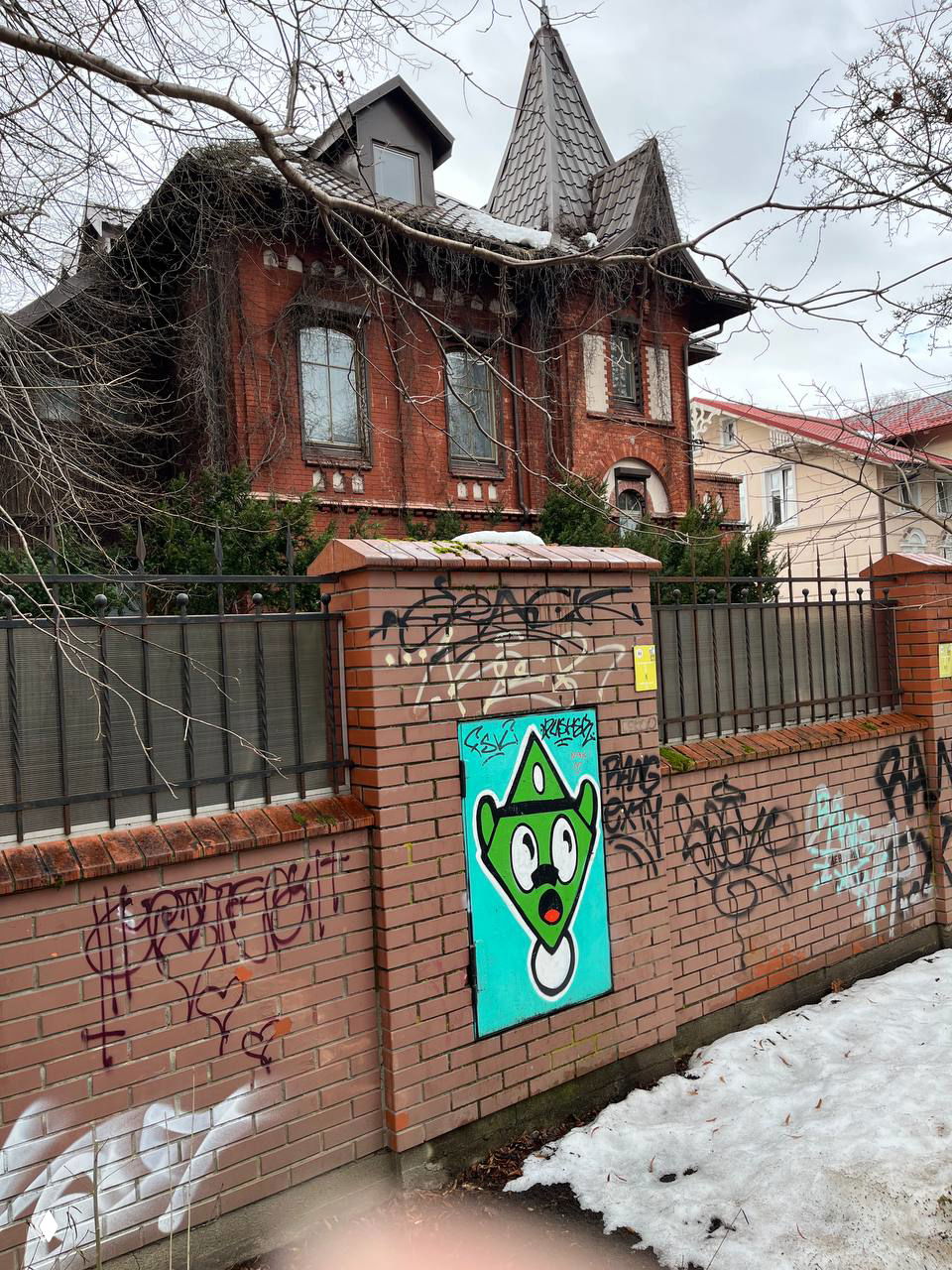 Особняк из красного кирпича с высокой шатровой крышей и декоративными элементами за кирпичным забором, на переднем плане видны граффити на стене.