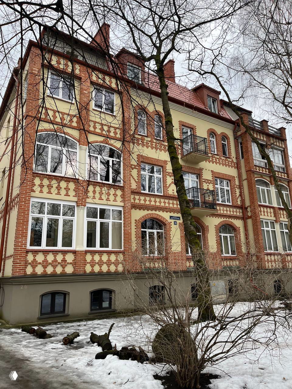 Кирпичный жилой дом с арочным входом, белыми архитектурными вставками и невысоким цоколем; снежные остатки в переднем плане и классические детали фасада.