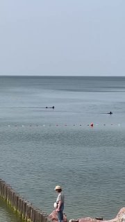 Спокойная гладь Балтийского моря с буйками и лодкой на горизонте; на переднем плане человек в шляпе идёт по берегу рядом с деревянными волнорезами.