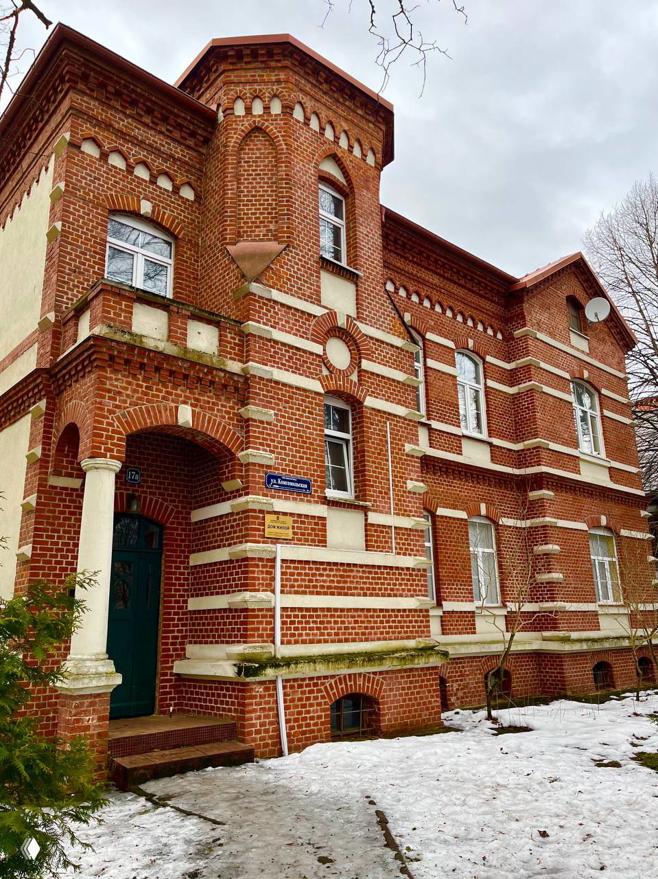 Невысокий кирпичный дом с башенкой и стрельчатыми мотивами в архитектуре конца XIX — начала XX века на ул. Комсомольской; сугробы и летние следы снега.