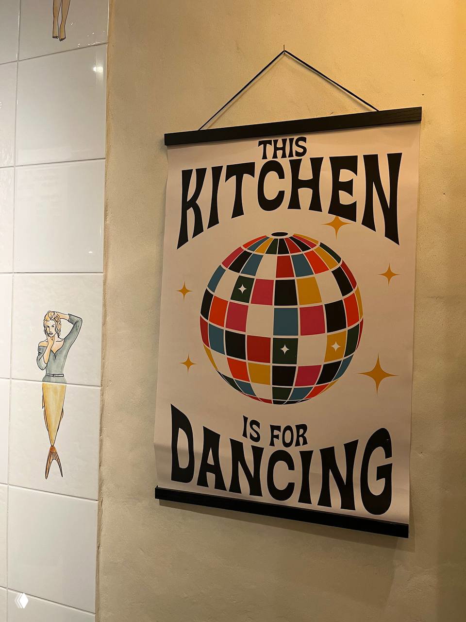 Плакат в интерьере кафе с надписью «THIS KITCHEN IS FOR DANCING» и цветным диско-шаром на фоне стены — часть декора заведения.
