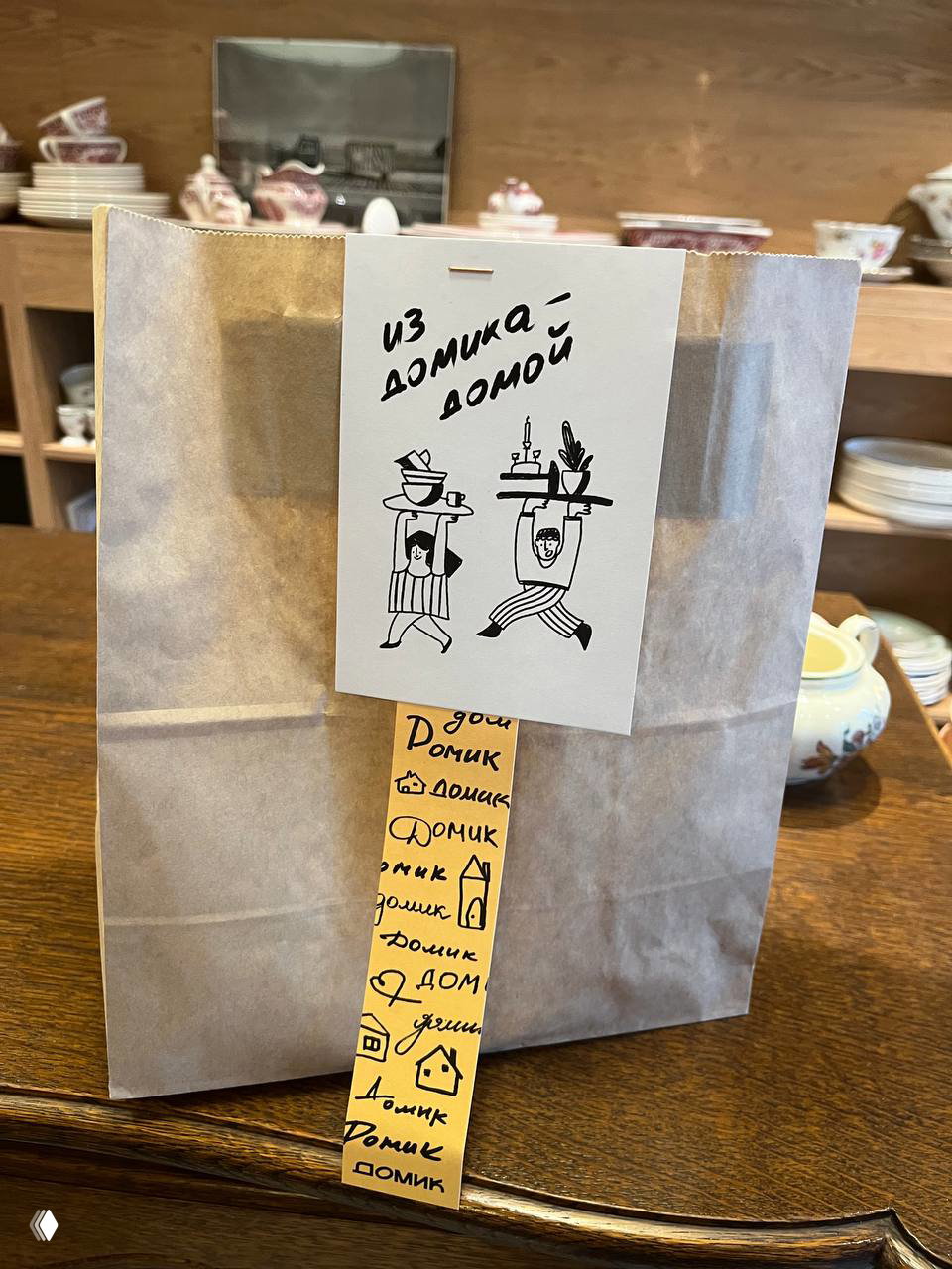 Бумажный пакет с яркой наклейкой и биркой «из домика домом» на прилавке — аккуратная упаковка покупки из кафе‑магазина.