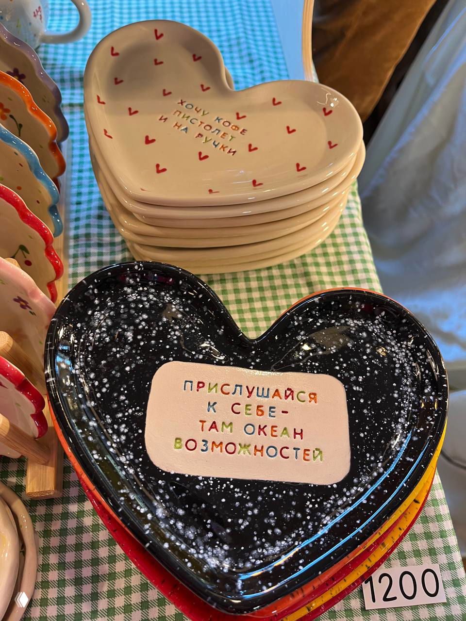 Сердцевидные керамические тарелки с надписями и разноцветной глазурью, выставленные на клетчатой ткани; виден ценник 1200 рублей.