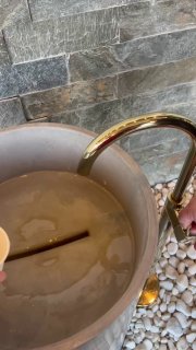 Крупный план чаши бювета с золотистым краном и минеральной водой; рядом виден бумажный стаканчик и струя воды из крана.