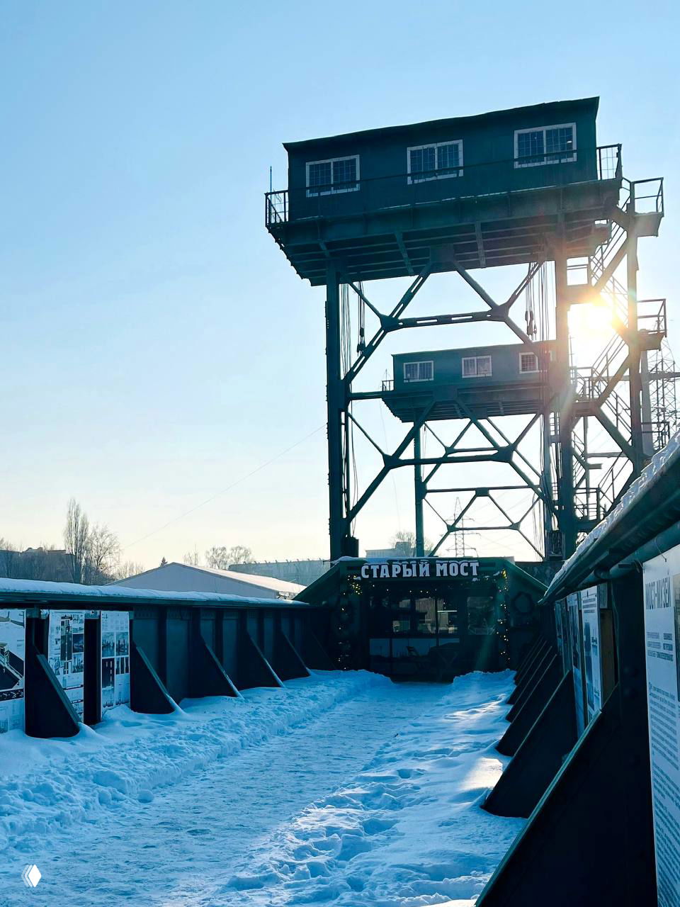 Металлическая опора старого моста с пешеходной дорожкой и выставочными стендами, снег и солнце создают контрастную зимнюю сцену.