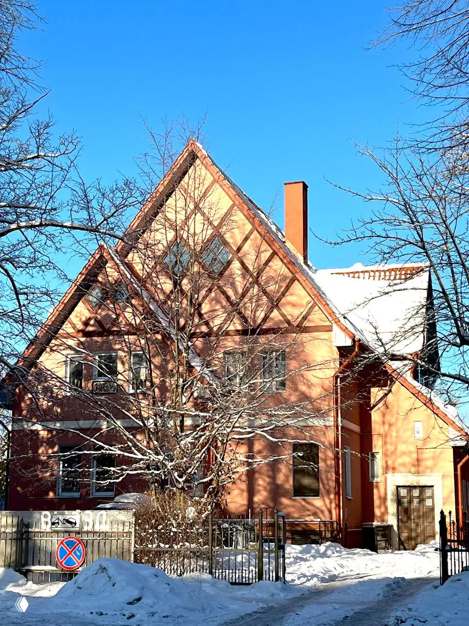 Красный кирпичный дом с высоким треугольным фронтоном и заснеженными деревьями перед фасадом — пример скромных вилл Марауненхофа на солнечном фоне.
