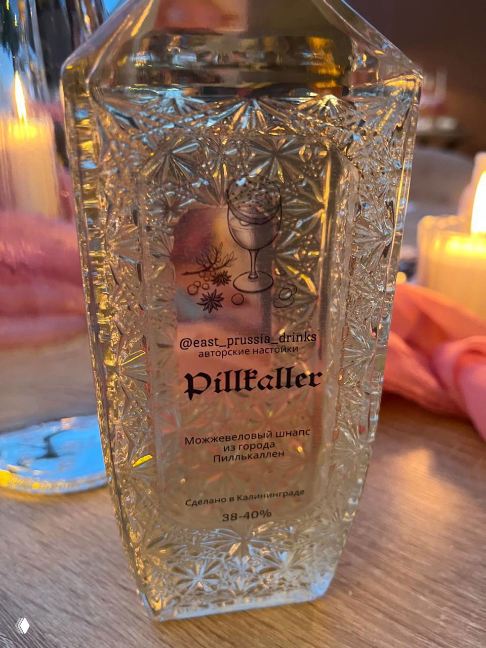 Крупный план декоративной стеклянной бутылки с этикеткой @east_prussia_drinks — напиток Pillkaller с текстовой информацией и узорчатым стеклом.