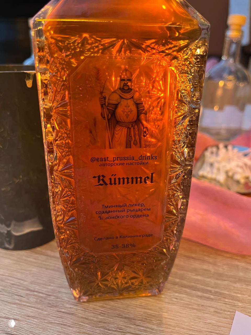 Стеклянная бутылка с коричневой настойкой и этикеткой Kummel; видна тёмная жидкость и рельефное декоративное стекло на столе мастерской.