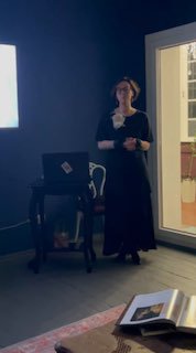 Лекторша в тёмном зале во время выступления; виден проекционный экран и уютное пространство арт‑квартиры, кадр вертикальный, немного смазан.