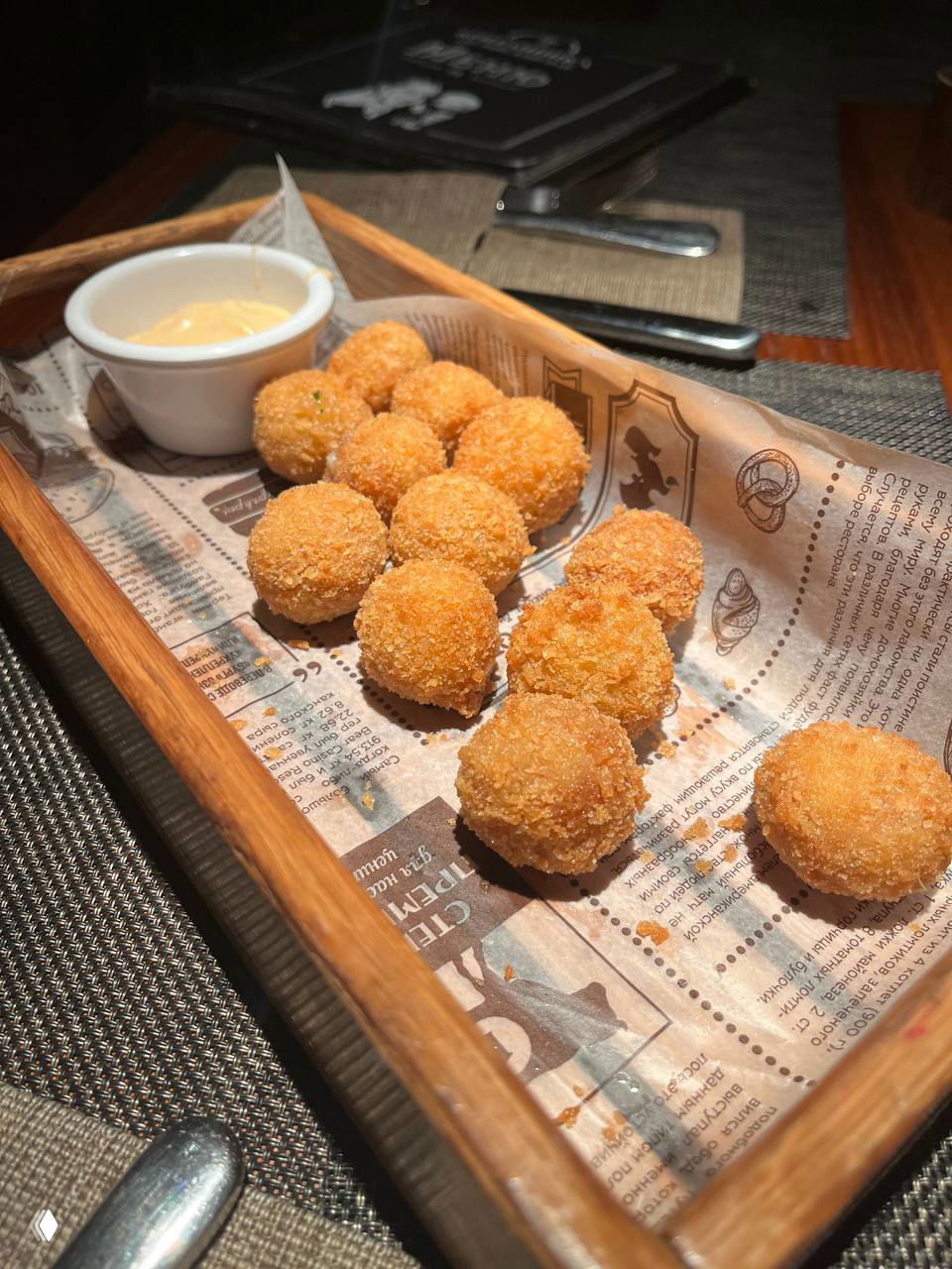 Небольшая деревянная корзинка с золотистыми сырными шариками и соусом для макания — закуска к пиву, сервированная на газетной бумаге.