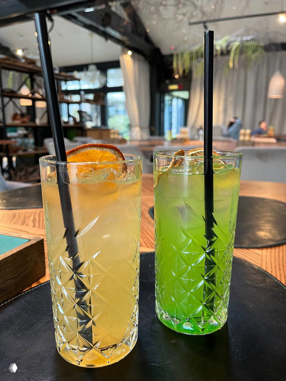Два высоких коктейля в гранёных стаканах с задекорированными дольками цитрусовых; вид на интерьер ресторана в глубине кадра.