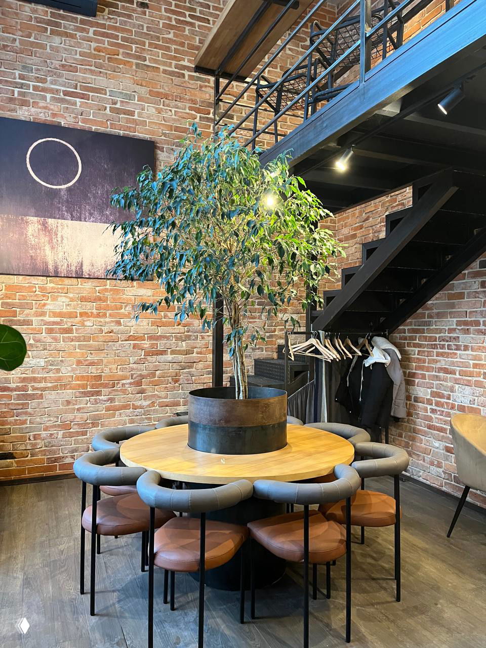 Интерьер кофейни «Символ»: круглый деревянный стол с растением в центре, вокруг низкие стулья, металлическая лестница и кирпичные стены.